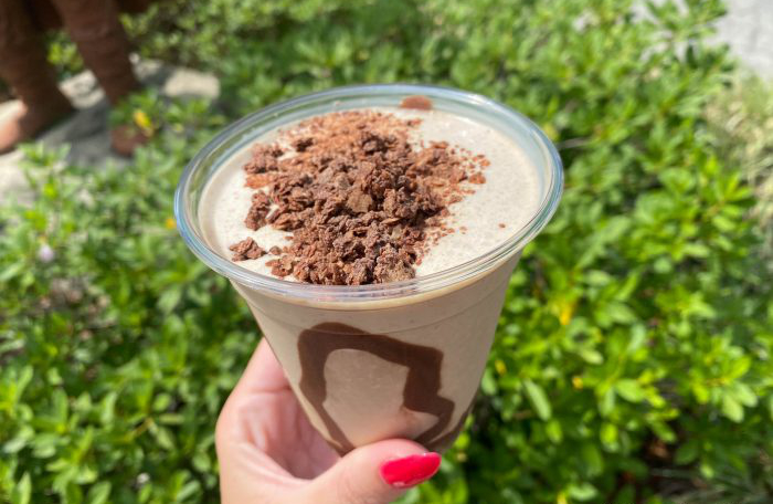 A plastic cup of viking coffee being held infront of bushes. Brown drink with chocolate sprinkles on top and chocolate sauce visible through cup.