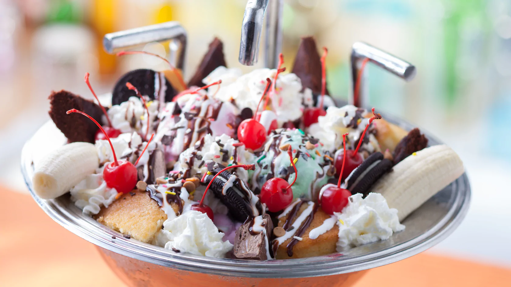 A picture of a huge bowl of ice cream, cherries, banana, cookies, cream, sauce, cake and chocolate chips with three straws