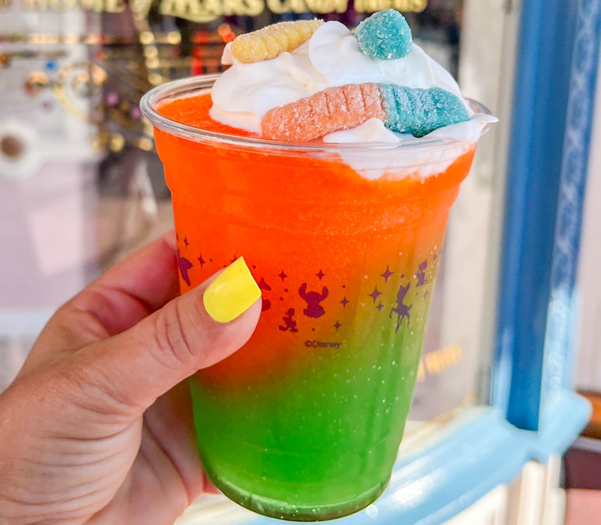 A plastic cup of the orange brrr'd being held infront of a shop window. The drink is orange and green coloured with whipped cream and gummy worms on top.