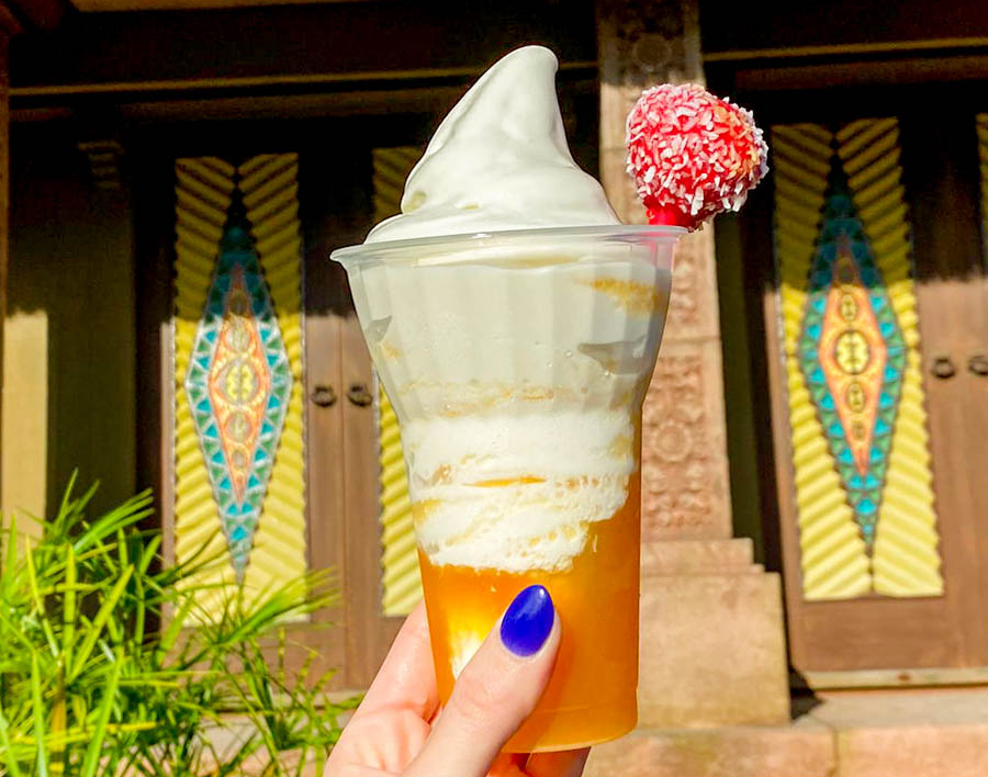 Picture of a tropical serenade drink with creamy dole whip on an orange juice base. The drink is being held up infront of Aloha Isle.